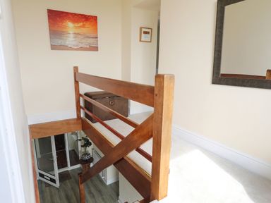 A staircase with wooden railing leading to an upper landing with a storage ottoman and wall art at Pant yr Hyman in Rhoscolyn