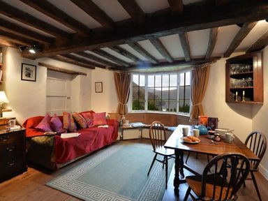 A living room with a red-covered sofa with cushions a wooden dining table with chairs and a bay window with curtains at Pantlleni Nant Gwynant