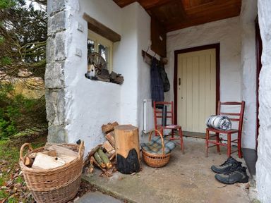 A porch with two chairs a basket with firewood a pile of logs a rolled blanket and a pair of boots at Pantlleni in Nant Gwynant