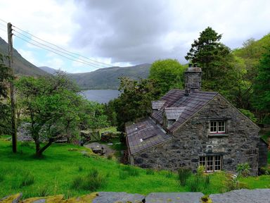 A stone house with a slate roof in a green rural landscape with trees hills and a lake at Pantlleni in Nant Gwynant