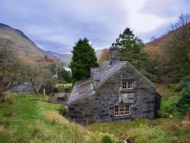 A stone cottage with a slate roof surrounded by trees and hills at Pantlleni in Nant Gwynant