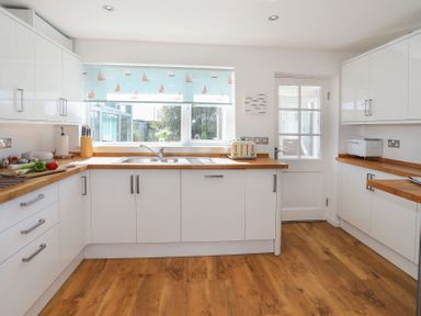 A kitchen with white cabinets wooden countertops a window with a sailboat blind a toaster and cutting board with vegetables at Pencraig in Trearddur Bay