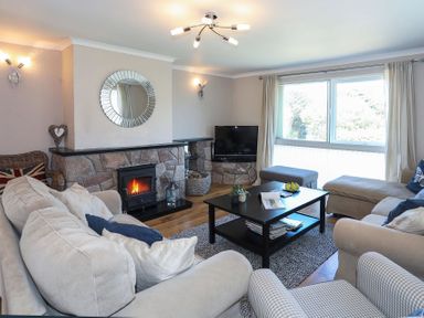 A living room with a stone fireplace a round mirror a television beige sofas a black coffee table and large window at Pencraig in Trearddur Bay