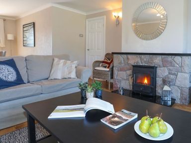 A living room with a sofa pillows a coffee table with books and pears a stone fireplace with a fire and a round mirror on the wall at Pencraig in Trearddur Bay