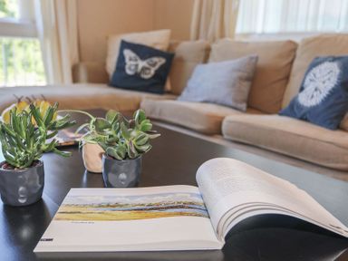 A coffee table with plants and an open book in front of a beige sofa with cushions at Pencraig in Trearddur Bay