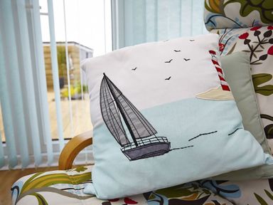 A cushion with a sailboat and lighthouse design on a floral armchair near a window at Pencraig in Trearddur Bay