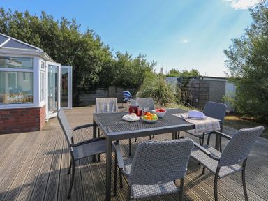 An outdoor wooden deck with a metal table and six woven chairs with fruit and drinks on the table at Pencraig in Trearddur Bay