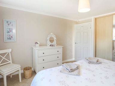 A bedroom with a bed towels on it a white chest of drawers with a decorative mirror a chair and a wicker basket at Pencraig in Trearddur Bay