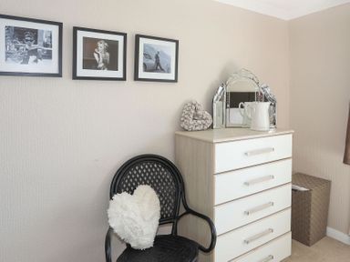 A black chair with a heart-shaped cushion next to a chest of drawers with a decorative mirror and pitcher on top and three framed photos on the wall at Pencraig in Trearddur Bay