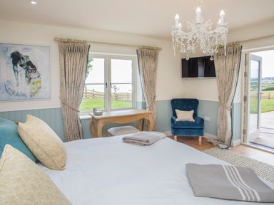 A bedroom with a bed pillows a blue armchair a desk a wall-mounted tv and an open door leading outside at Penrhyn Barn in Dulas