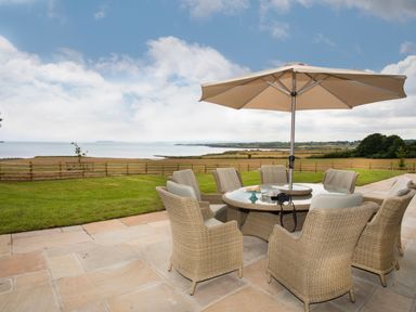 An outdoor patio with wicker chairs around a table and an umbrella overlooking a fenced grassy area and water at Penrhyn Barn in Dulas