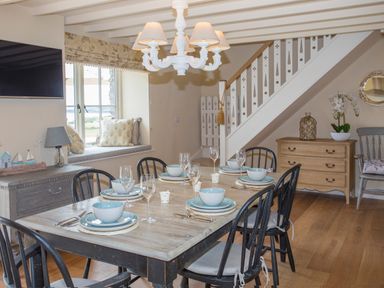 A dining room with a wooden table set for six under a chandelier next to a window seat and staircase at Penrhyn Barn in Dulas