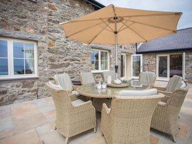 An outdoor patio with a round glass table surrounded by wicker chairs and a large beige umbrella at Penrhyn Barn in Dulas
