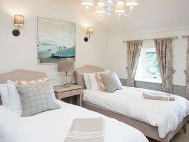 A bedroom with two single beds bedside table lamp wall lights painting and curtained window at Penrhyn Barn in Dulas