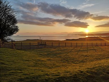 A grassy field with a wooden fence overlooking the sea at sunset at Penrhyn Barn in Dulas