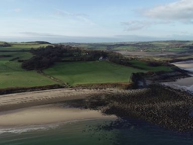 A coastal area with sandy and rocky shorelines and green fields with scattered trees and buildings at Penrhyn Barn in Dulas