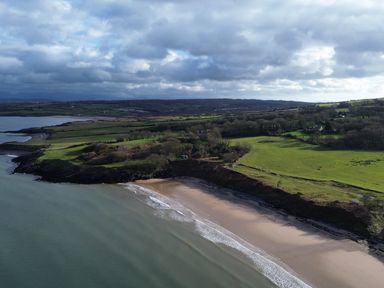 A coastal landscape with sandy beach green fields and scattered trees under a cloudy sky at Penrhyn Barn in Dulas