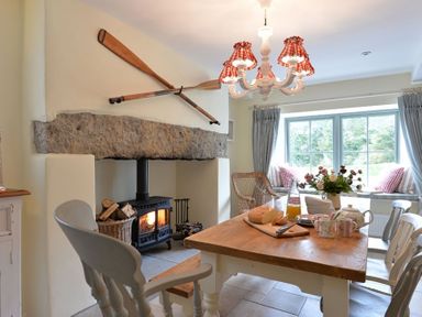 A dining area with a wooden table set with mugs bread and flowers near a wood stove with oars mounted above at Penrhyn Farm in Moelfre