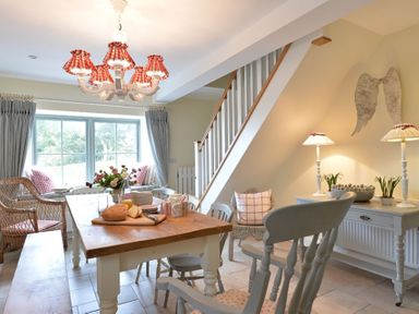 A dining area with a wooden table, chairs, a bench, flowers, and a wicker armchair near a window at Penrhyn Farm in Moelfre