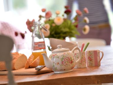 A table with a floral teapot, cups, sliced bread, a bottle of orange juice, and flowers in the background at Penrhyn Farm in Moelfre