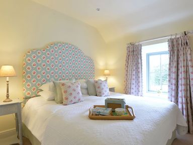 A bedroom with a large bed with floral headboard and pillows bedside tables with lamps and a window with floral curtains at Penrhyn Farm Moelfre