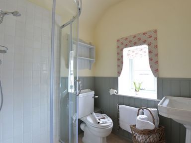 A bathroom with a glass shower, toilet, sink, window with floral curtain and towel basket at Penrhyn Farm in Moelfre