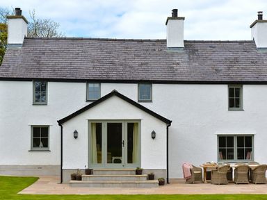 The back of a white two-story house with a slate roof a patio with wicker chairs and a table and steps leading to glass doors at Penrhyn Farm in Moelfre