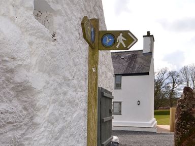 A walking direction sign attached to a wooden post next to a white stone building with a house in the background at Penrhyn Farm in Moelfre