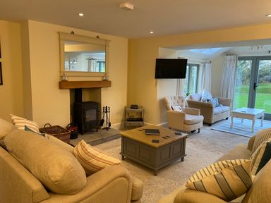 A living room with beige sofas, a coffee table, a wood-burning stove, an armchair, a wall-mounted television, a wicker sofa, and glass doors leading to a garden at Penrhyn Farm in Moelfre