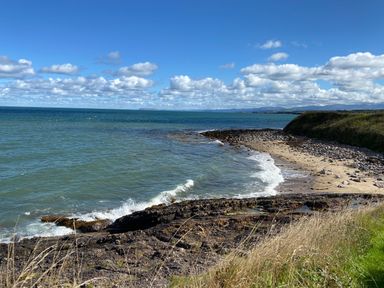 A rocky shoreline with grass in the foreground and the sea under a blue sky at Penrhyn Farm in Moelfre