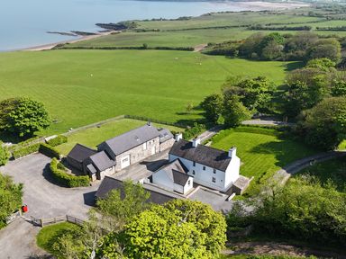 An aerial view of a farmhouse with multiple buildings surrounded by green fields and trees near a coastline at Penrhyn Farm in Moelfre