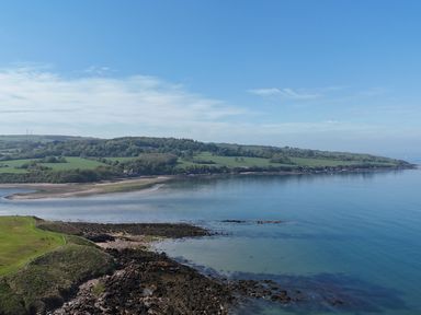 A coastal landscape with rocky shoreline green fields and wooded hills under a blue sky at Penrhyn Farm in Moelfre