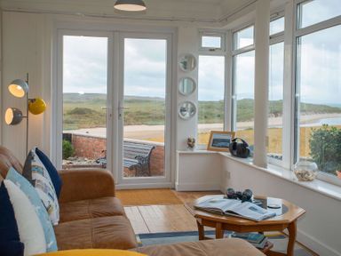 A sitting area with a brown couch pillows a wooden coffee table and windows overlooking a beach at Pen Y Daith in Aberffraw