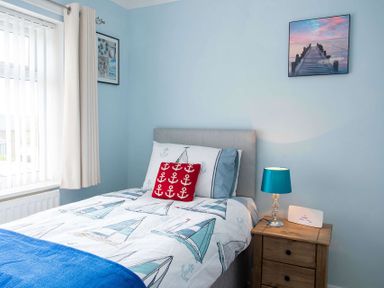 A bedroom with a bed with boat patterned bedding a bedside table with a lamp and framed pictures on blue walls at Pen Y Daith in Aberffraw