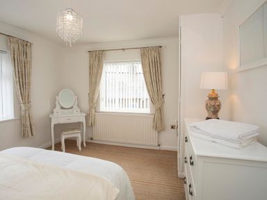 A bedroom with a white dressing table and stool a window with curtains and a white chest of drawers with a lamp and towels at Pen Y Daith in Aberffraw
