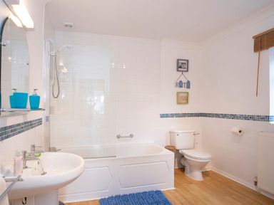 A bathroom with a bathtub and shower glass screen a white toilet and sink with blue accessories and wooden floor at Pen Y Daith in Aberffraw