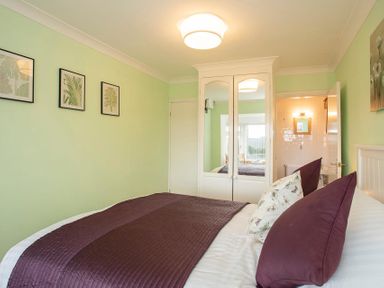 A bedroom with a bed covered in white and purple linens a white wardrobe with mirrored doors and framed botanical prints on a green wall at Pen Y Daith in Aberffraw