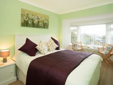 A bedroom with a white bed and purple bedding next to a window with two chairs and a small table at Pen Y Daith in Aberffraw
