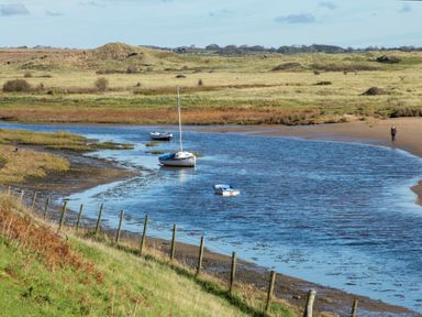 A river with boats moored near grassy banks and a person walking on a sandy shore at Pen Y Daith in Aberffraw