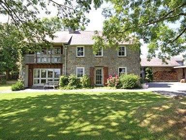 A stone house with wooden shutters and balcony surrounded by green lawn and trees at Perth Hwfa in Malltraeth
