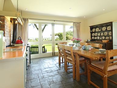 A kitchen dining area with wooden table and chairs set with dishes and flowers near large windows at Perth Hwfa in Malltraeth