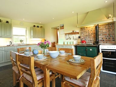 A kitchen with a wooden dining table set with striped bowls and plates next to a green stove and white oven at Perth Hwfa in Malltraeth