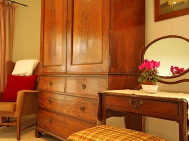 A room with a wooden wardrobe a wooden dressing table with a round mirror a chair with cushions and a potted plant on the table at Perth Hwfa in Malltraeth