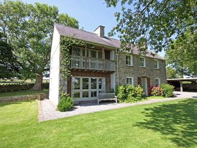 A two-story stone house with a balcony and garden surrounded by trees at Perth Hwfa in Malltraeth