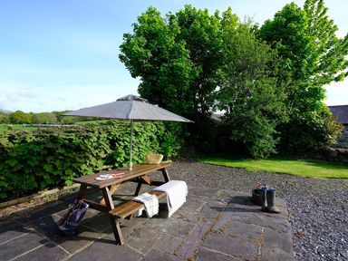 A wooden picnic table with an umbrella on a stone patio near trees and grass at Pen Y Bont in Pontllyfni