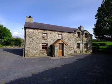 A stone house with a wooden door and multiple windows surrounded by gravel and trees at Pen Y Bont in Pontllyfni