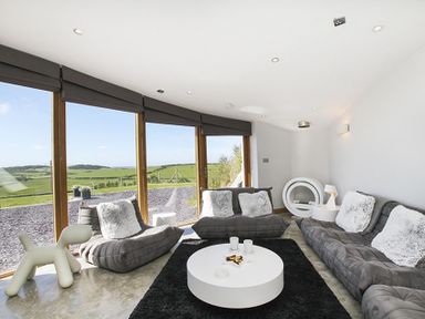 A living room with gray sofas and cushions a round white coffee table black rug and large windows showing green fields at Rhyd Angharad Barns in Rhydwyn