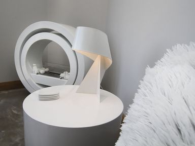 A round white side table with a modern white lamp and coasters beside a white fluffy chair and circular fireplace in Rhyd Angharad Barns in Rhydwyn
