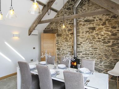 A dining room with a white table and six chairs set with plates and glassware next to a stone wall with a wood burning stove at Rhyd Angharad Barns in Rhydwyn