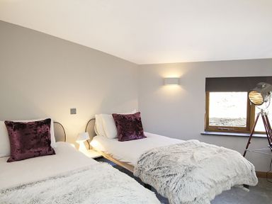 A bedroom with two single beds white bedding purple pillows a window and a tripod floor lamp at Rhyd Angharad Barns in Rhydwyn
